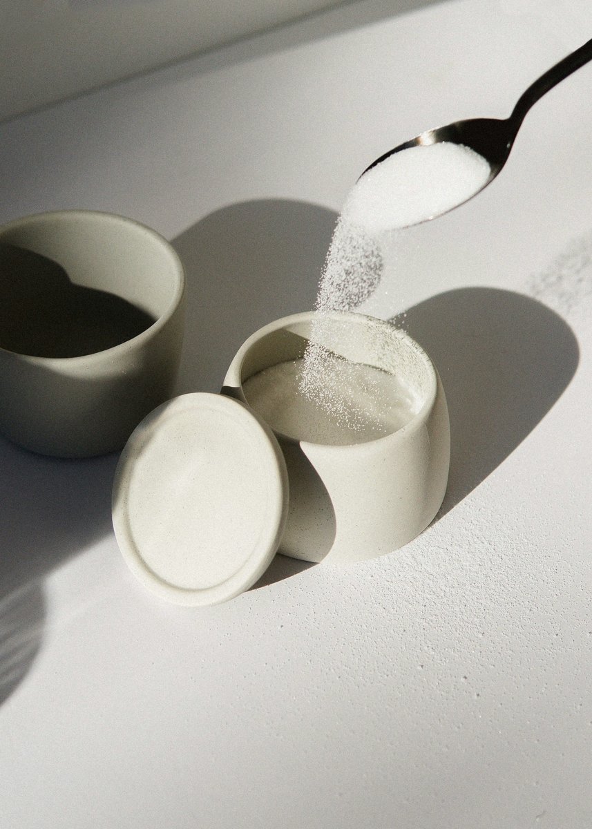 Cuillère de sucre versée dans un sucrier blanc sous une lumière rasante